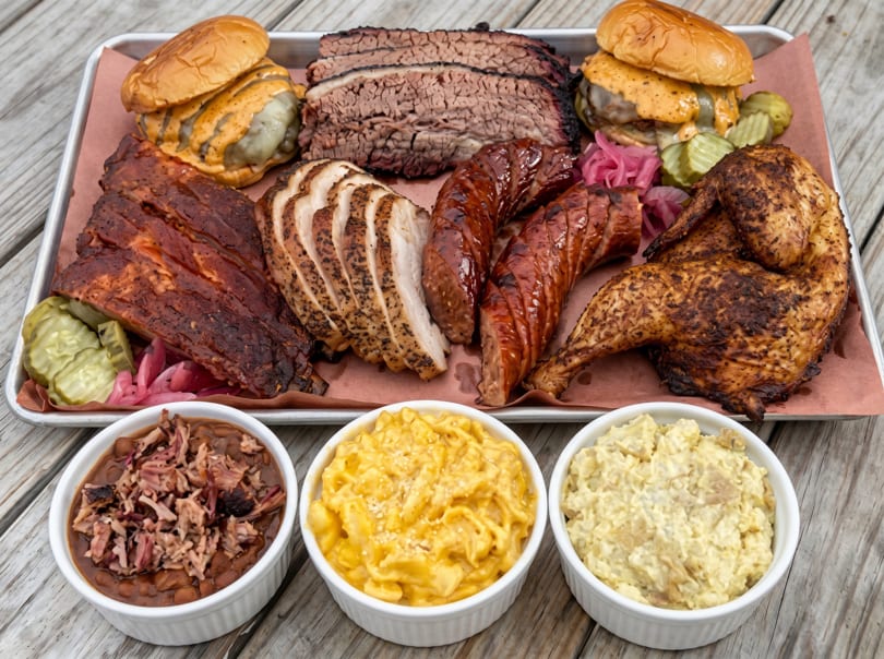 A large platter of barbecue meats including brisket, ribs, chicken, sausages, and sandwiches with sides.