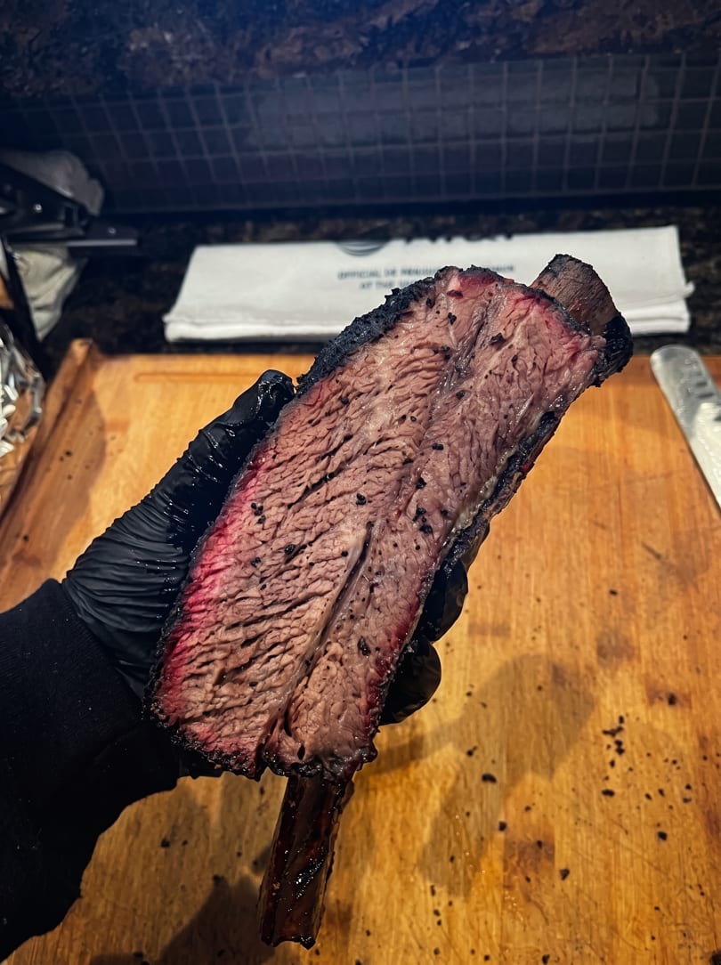 A gloved hand holds up a large, perfectly smoked beef rib with a prominent smoke ring.